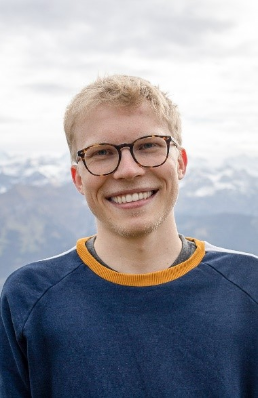 Photo de Janosch Herzog. M. Herzog porte des lunettes et sourit à la caméra. On ne voit que le haut de son corps. Il porte un pull-over bleu. En arrière-plan, on voit des montagnes et un ciel nuageux. 